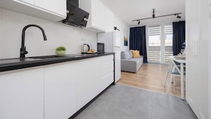 A modern kitchen with white cabinets, a black faucet, and a small island. There is a dining area with a table and chairs, a gray sofa, and a view of the outside through large windows with blue curtains.