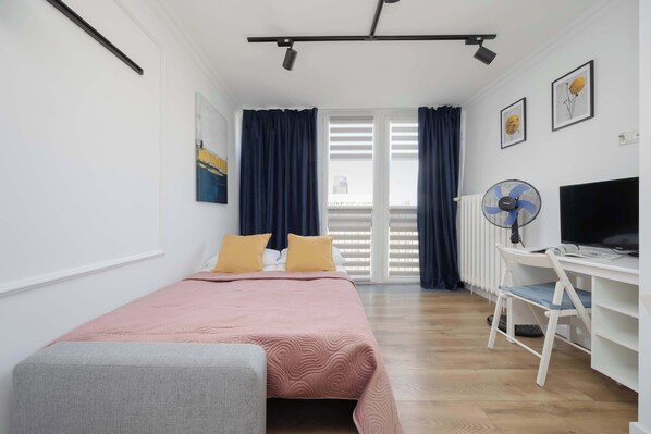 Une chambre à coucher avec un lit, un bureau et une chaise. Il y a une fenêtre avec des rideaux, un radiateur et un ventilateur fixé au mur.