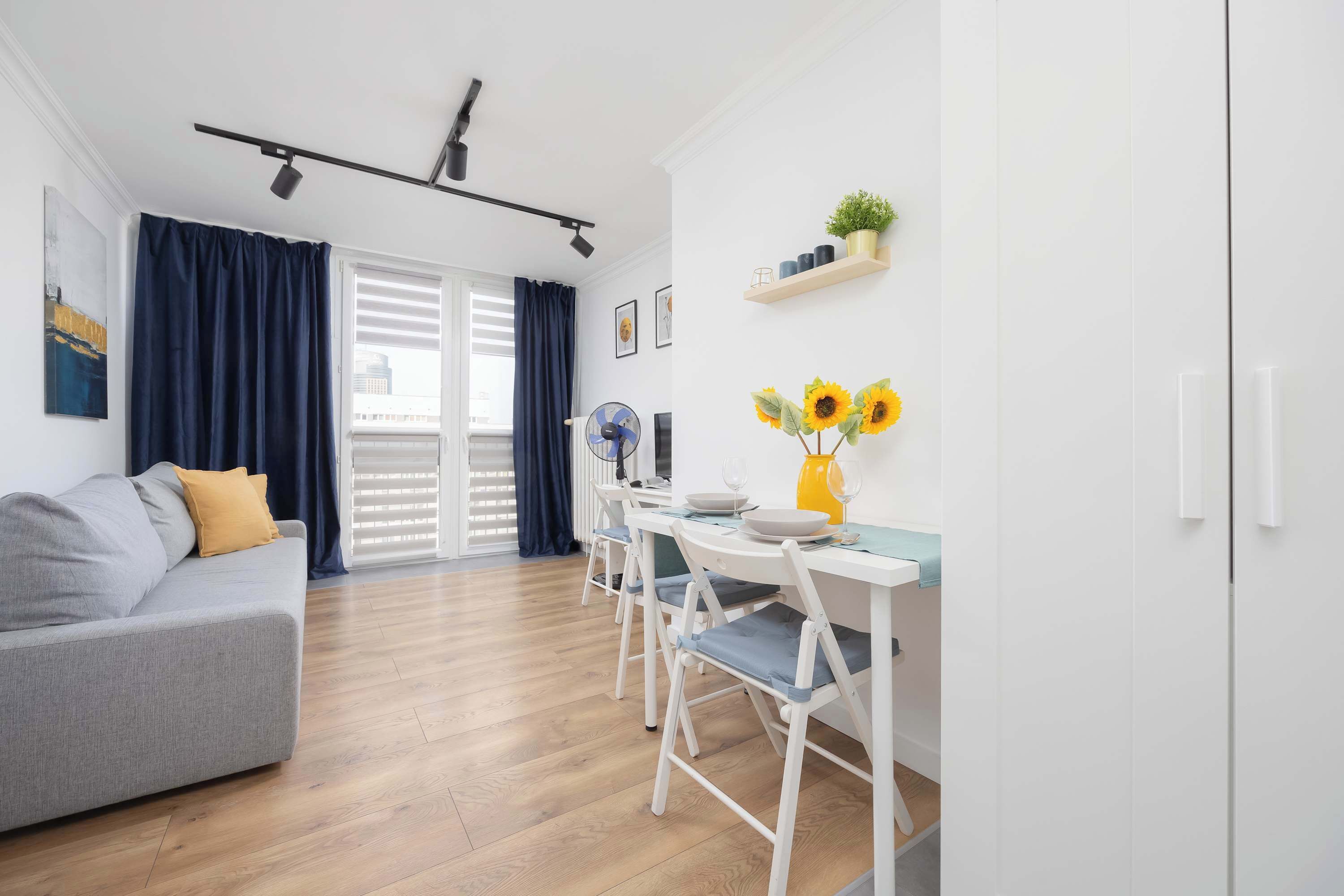 Un salon moderne avec un canapé gris, une table à manger avec des chaises, un luminaire au plafond et une fenêtre avec des rideaux.
