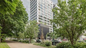 Un bâtiment moderne à la façade en briques et aux grandes fenêtres, entouré d’une végétation luxuriante et bordé d’un chemin pavé.