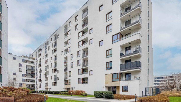 Un edificio residencial de varios pisos con balcones, un sendero pavimentado y zonas ajardinadas.