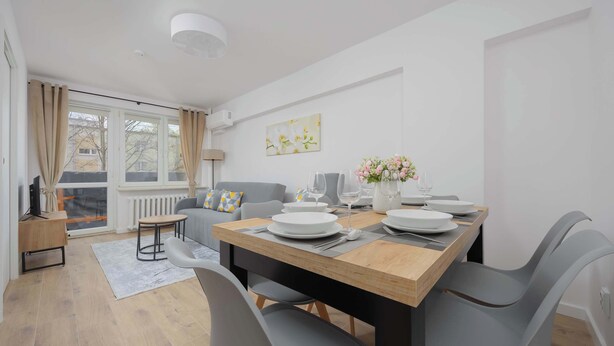 Un espace repas moderne avec une table en bois dressée pour quatre personnes, entourée de chaises grises. Un canapé gris et une petite table d’appoint avec une lampe sont visibles à l’arrière-plan.