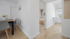 Une pièce moderne et bien éclairée, avec un coin repas comprenant une table en bois et des chaises grises. On aperçoit à l’arrière-plan un lit recouvert d’une couverture beige et une fenêtre avec des rideaux.