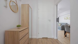 Un couloir avec une commode en bois, un miroir et une porte donnant sur un coin repas avec un canapé gris et une table.