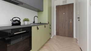 A modern kitchen with a black stove, a kettle, and a sink. There is a wooden door and a white wall in the background. - Stylish Studio with Air Conditioning | Close to EXPO XXI (Warszawa)