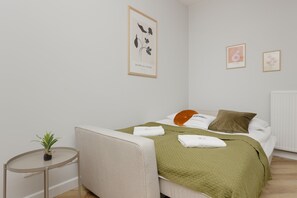 Ein ordentlich eingerichtetes Schlafzimmer mit einem Bett, Kissen, einer grünen Decke, einem kleinen Tisch mit einer Pflanze und gerahmten Kunstwerken an der Wand.
