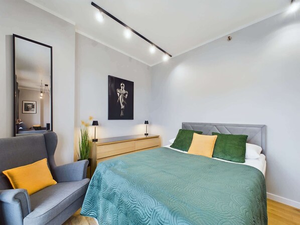 Un dormitorio moderno con una cama, un sofá gris, una mesita de noche, un espejo y una obra de arte enmarcada en la pared.