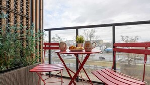 Ein Balkon mit roten Metallstühlen, einem kleinen Tisch, einem Blumentopf und Blick auf die Stadt.