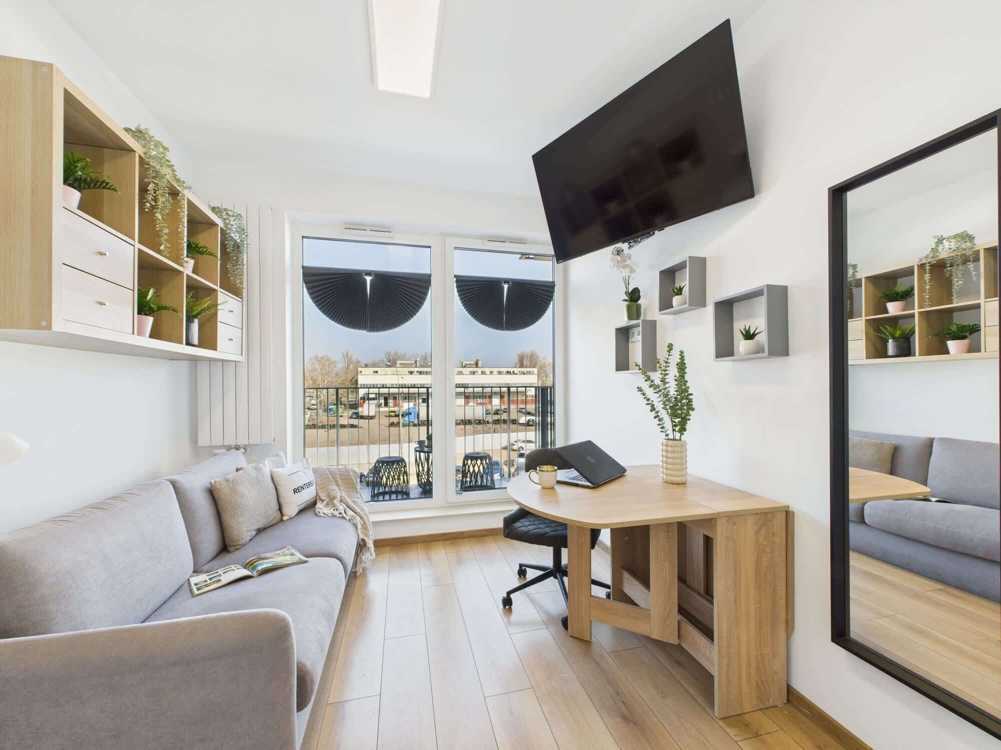 Un salon moderne avec un canapé, un bureau en bois, un téléviseur à écran plat et une grande fenêtre donnant sur la ville.