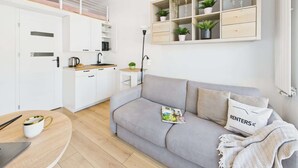 Un salon moderne avec un canapé gris, une table basse et un magazine. On y trouve également un coin cuisine avec des meubles blancs et une étagère suspendue pour les plantes.
