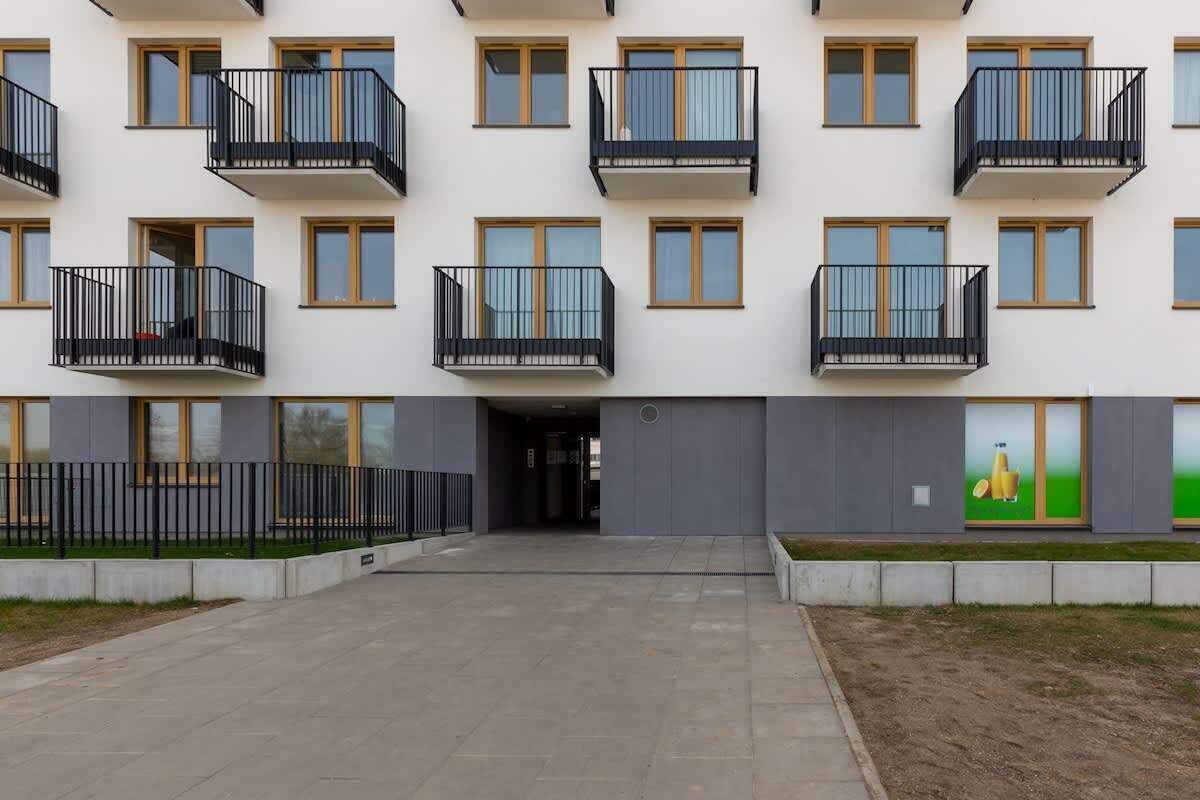 A multi-story residential building with balconies, a central entrance, and a landscaped area with a low fence.