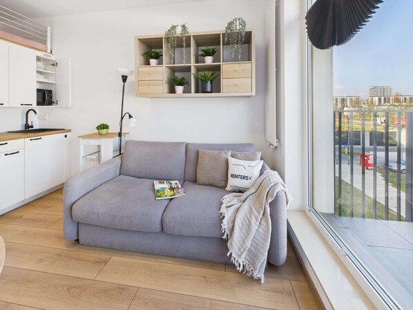 Een moderne woonkamer met een grijze bank, een tijdschrift en een 'RENTERS' kussen. Er is een houten plank met planten en een raam met uitzicht op gebouwen.