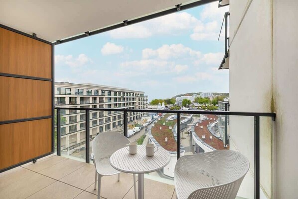 A balcony with two white metal chairs and a small table, overlooking a cityscape with modern buildings and a road.