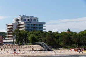 Ein modernes Hotel mit mehreren Etagen, Balkonen und einem Strandbereich, der über eine Treppe erreichbar ist.