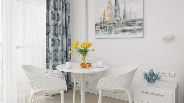 A modern room with a round table, two white chairs, a vase with yellow flowers, and a painting of sailboats on the wall.