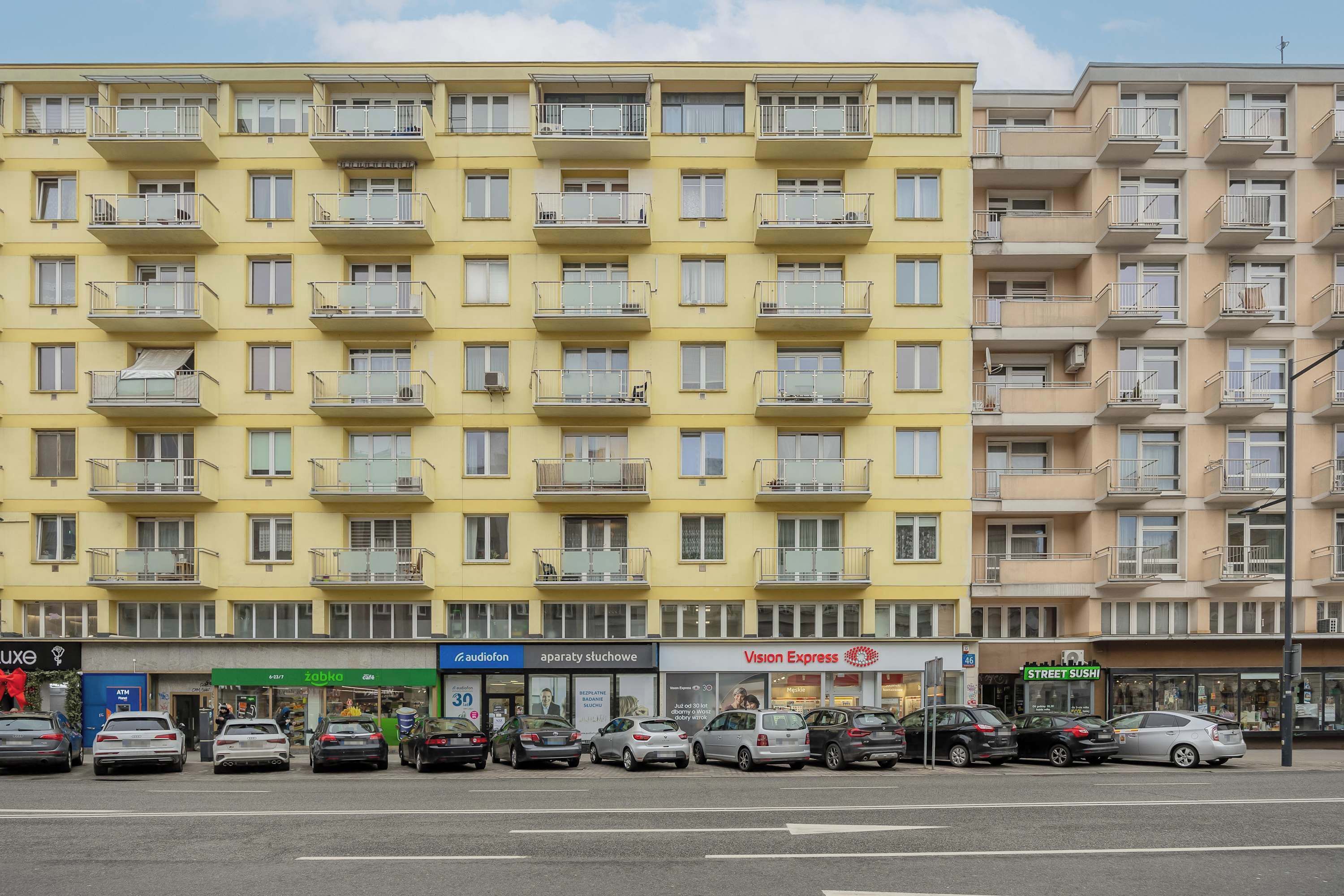 Ein mehrstöckiges Wohnhaus mit Balkonen, eine Reihe geparkter Autos und verschiedene Geschäfte, darunter eine Apotheke und ein Sushi-Restaurant.