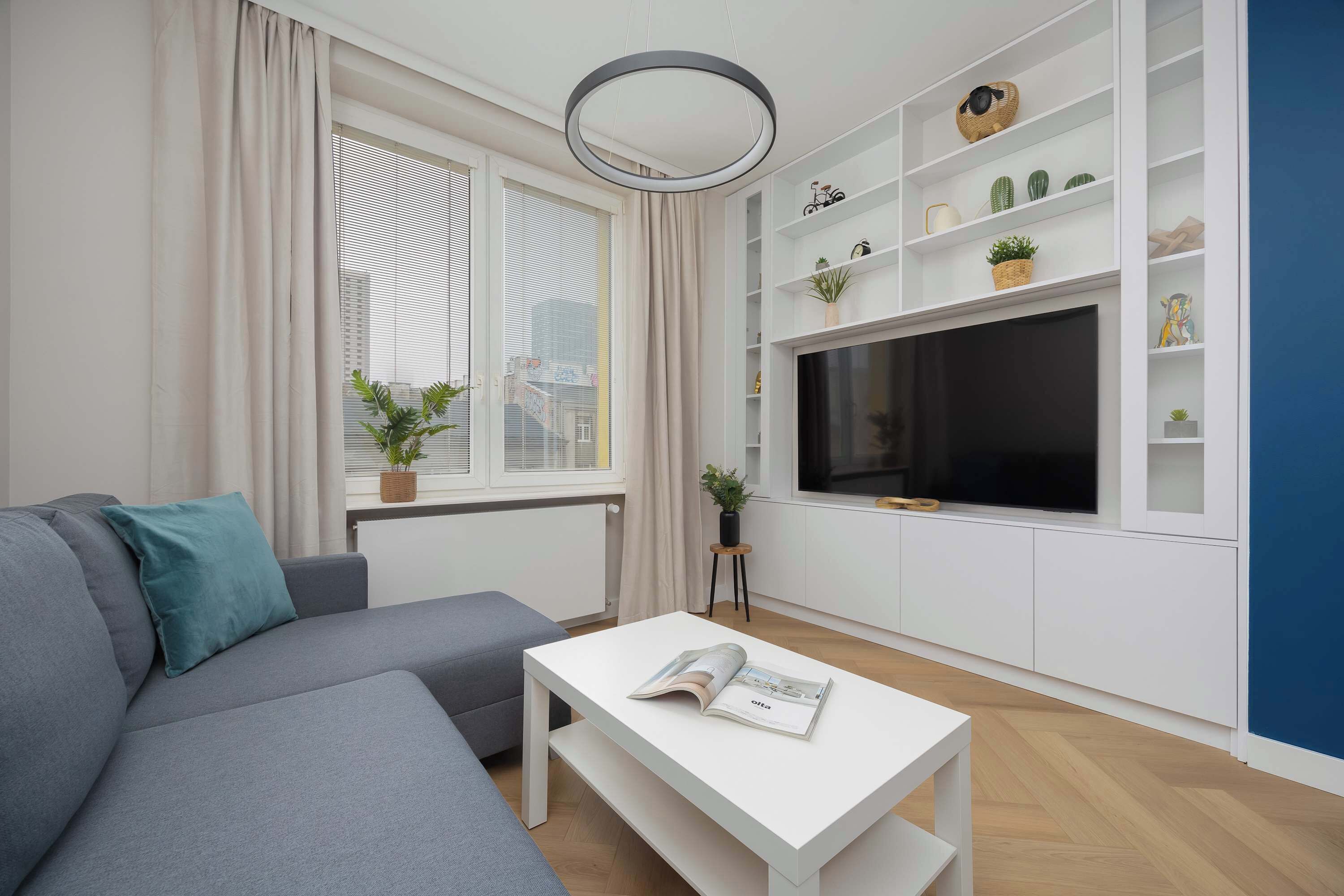 Ein modernes Wohnzimmer mit einem grauen Sofa, einem weißen Couchtisch mit einer Zeitschrift, einem Fernseher auf einem weißen Regal und einem Fenster mit Jalousien.