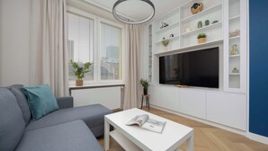 A modern living room with a grey sofa, a white coffee table with a magazine, a TV on a white shelf, and a window with blinds. - Stylish Apartment in the Center | Workplace (Warszawa)