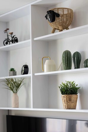 A modern interior with white shelves displaying various decorative items including a wicker basket with a black sheep figure, a small bicycle, and several potted plants.