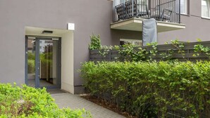 Un edificio moderno con fachada gris, puerta de entrada de vidrio, balcón con barandilla metálica y un seto verde al frente.