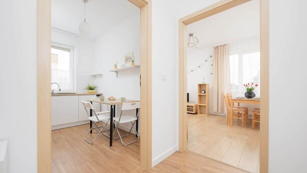 Una cocina moderna con armarios blancos, un comedor con mesa y sillas de madera, y vistas a través de un acceso abierto.
