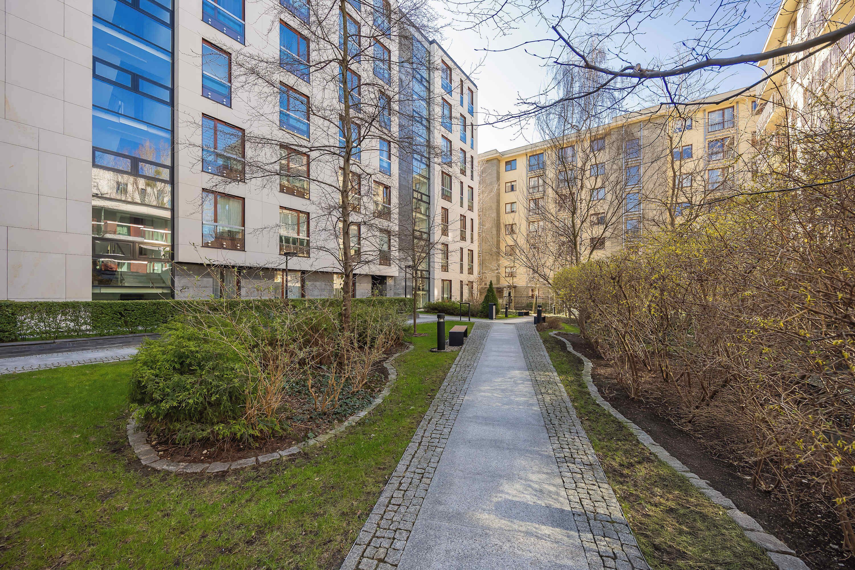 Un sendero bordeado por edificios modernos, un césped bien cuidado y un seto.
