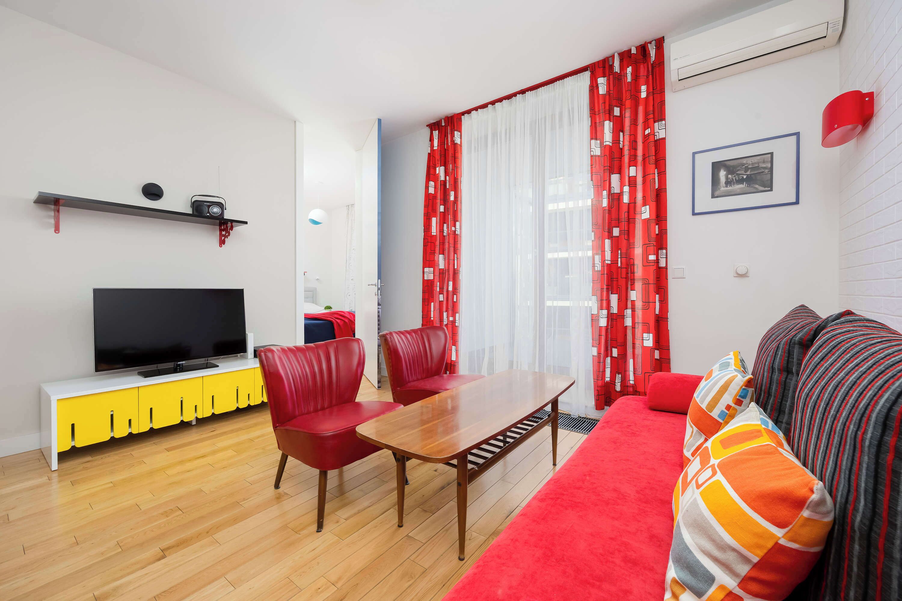 Una sala de estar moderna con un televisor de pantalla plana sobre un mueble de televisión amarillo y blanco, sillas rojas, una mesa de centro de madera y un sofá rojo con cojines a rayas.
