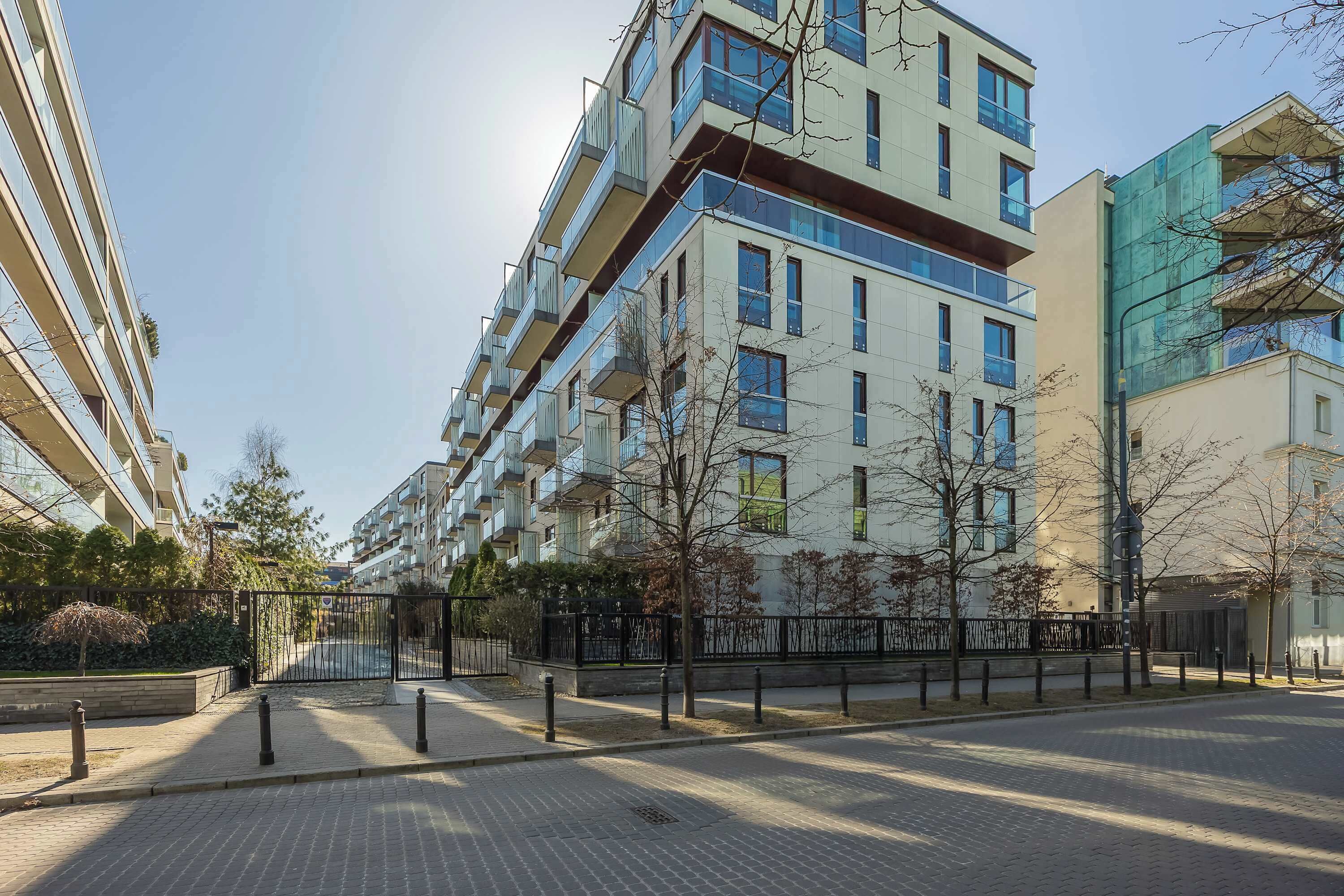 Un moderno edificio de apartamentos con varios balcones, entrada protegida por un portón y un camino pavimentado.