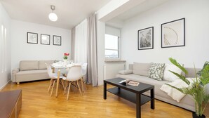 Una sala de estar moderna con sofá, mesa de comedor y sillas. Se observan cuadros en las paredes y una planta en maceta.