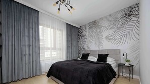 A modern bedroom with a large bed, grey curtains, a wall mural, and a bedside table with a lamp.