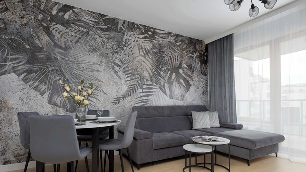 A modern living room with a grey sofa, two grey chairs, a round dining table, and a small side table. A large window with sheer curtains is visible.