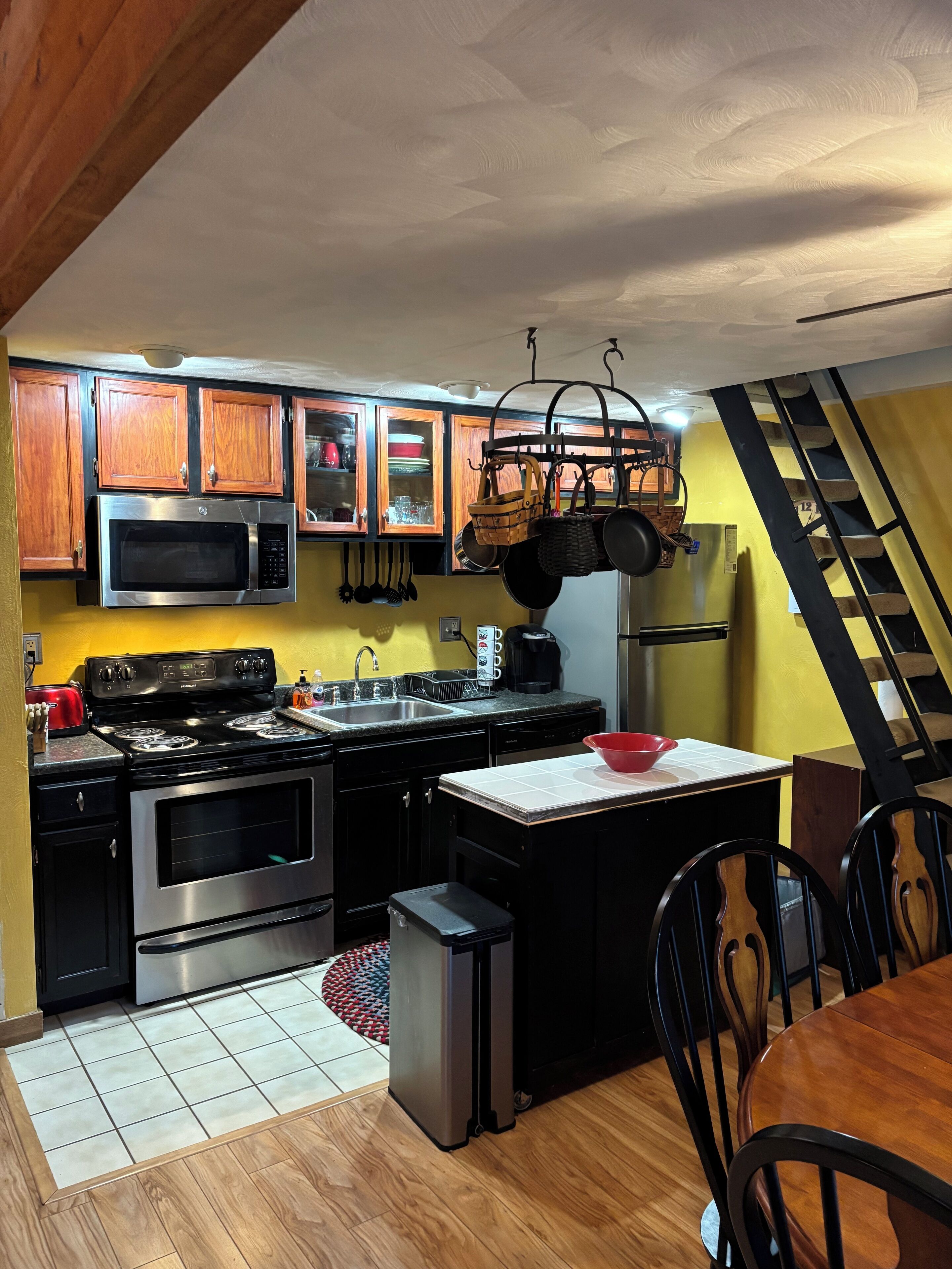 A kitchen with wooden cabinets, a microwave, stove, and a sink. There is a red bowl on the counter and a trash can.