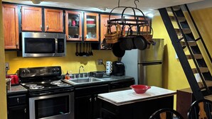 Een keuken met houten kastjes, een magnetron, fornuis en een spoelbak. Op het aanrecht staat een rode kom en er staat een prullenbak.