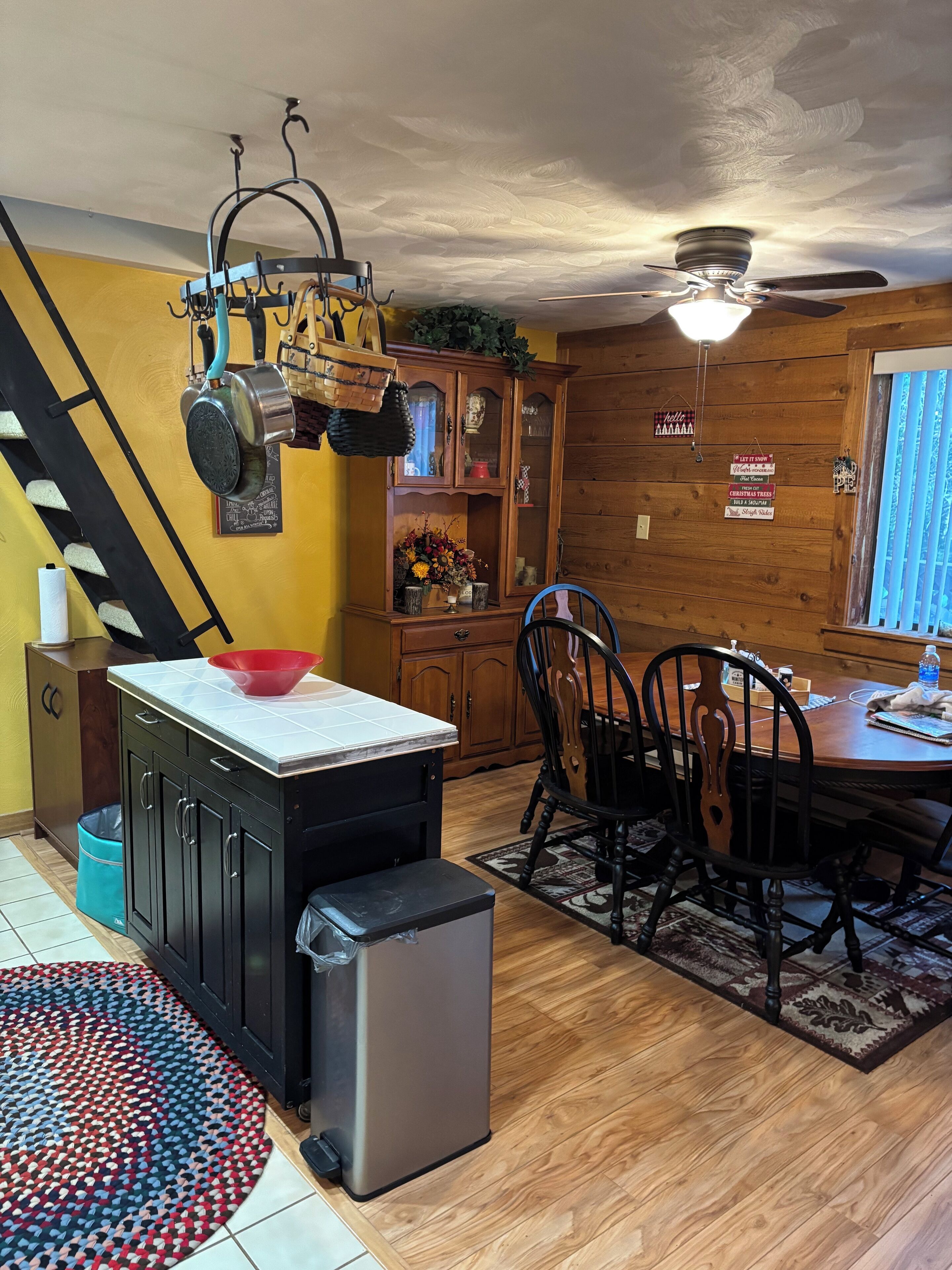 Una cocina con una isla central, un comedor con mesa y sillas de madera, un ventilador de techo y una escalera.
