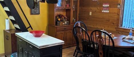 Una cocina con una isla central, un comedor con mesa y sillas de madera, un ventilador de techo y una escalera.