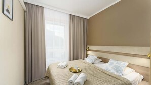 Una habitación de hotel con una cama, cortinas, un cabecero y una mesita de noche con una bandeja de objetos.
