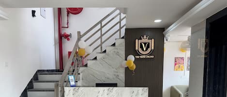 Uma área de recepção com balcão de mármore, escada com degraus em preto e branco e parede com logotipo e uma fotografia emoldurada.