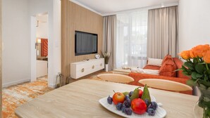 A modern hotel room with a wooden accent wall, a flat-screen TV, a dining area with a fruit plate, and a cozy orange sofa.