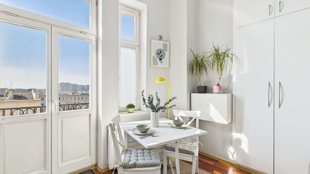 A modern kitchen with white cabinetry, a dining table with chairs, a window with a view of buildings, and a potted plant on a shelf. - Cozy Studio in the Center | Balcony and Proximity to Bathrooms (Warszawa)