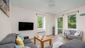 A modern living room with a grey sofa, a wooden coffee table, and a grey couch with striped cushions. There is a flat-screen TV mounted on the wall and a window with curtains.