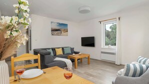 A modern living room with a sofa, a dining table with wine glasses, a TV mounted on the wall, and a window with curtains.