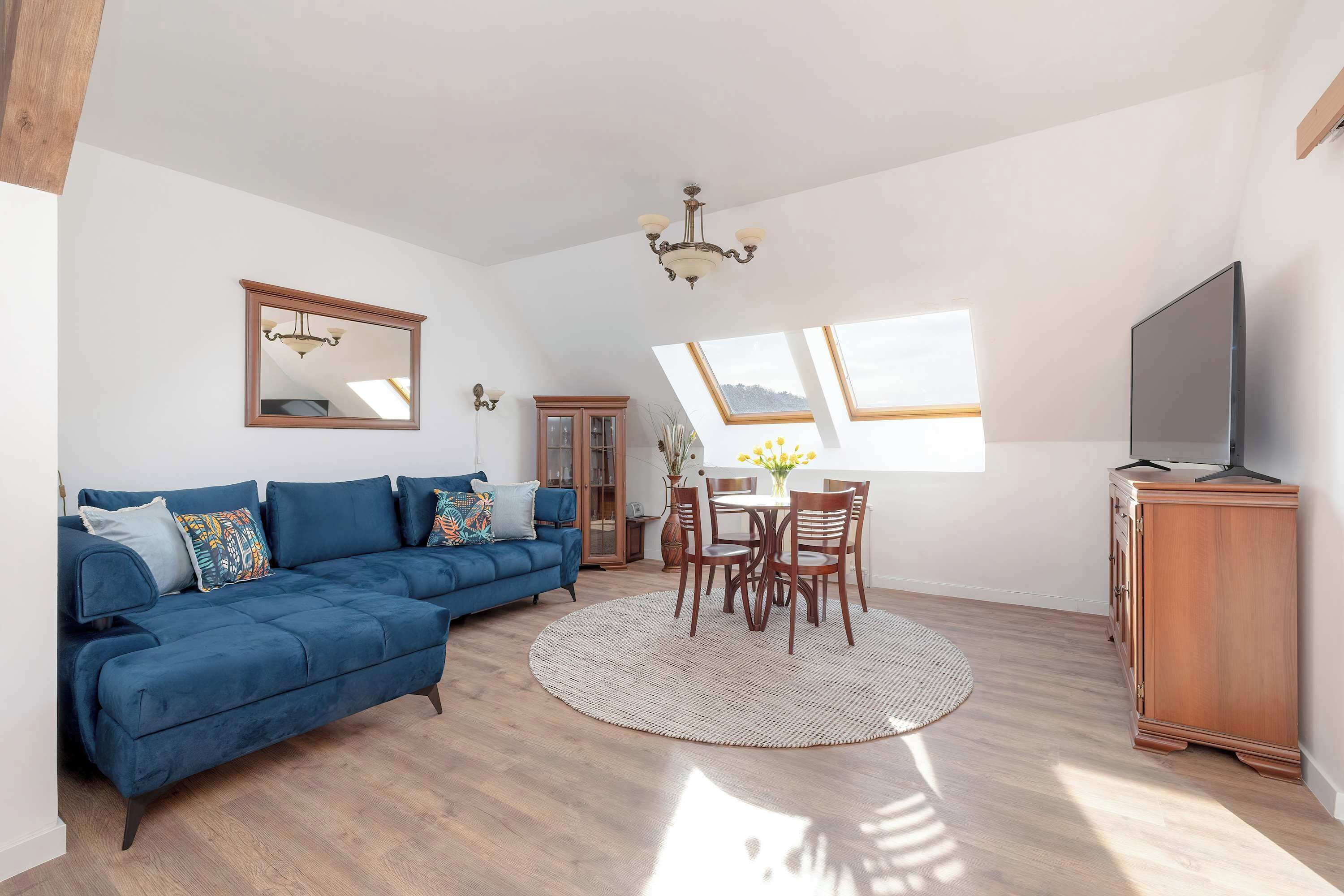 A spacious living area with a blue sofa, a dining table with chairs, a wooden cabinet, and a large mirror on the wall.