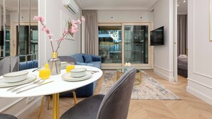 A modern living room with a dining table set for four, a blue sofa, and a large window with a view of the city.