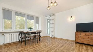 Una habitación amplia con piso de madera, una mesa de comedor con sillas, un televisor sobre un mueble de madera y un ventanal con vista a árboles.