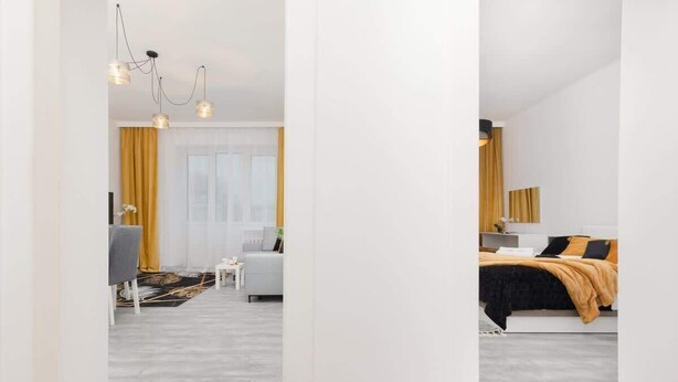 A modern hotel room with a grey floor, a bed with a black and orange bedspread, a white sofa, and a small table. There is a window with yellow curtains and a chandelier with three light fixtures. - Spacious Apartment | 2 Bedrooms | Workspace (Warszawa)