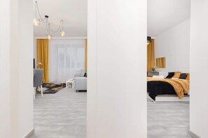 Une chambre d’hôtel moderne avec un sol gris, un lit recouvert d’une couvre-lit noire et orange, un canapé blanc et une petite table. Il y a une fenêtre avec des rideaux jaunes et un lustre à trois lampes.