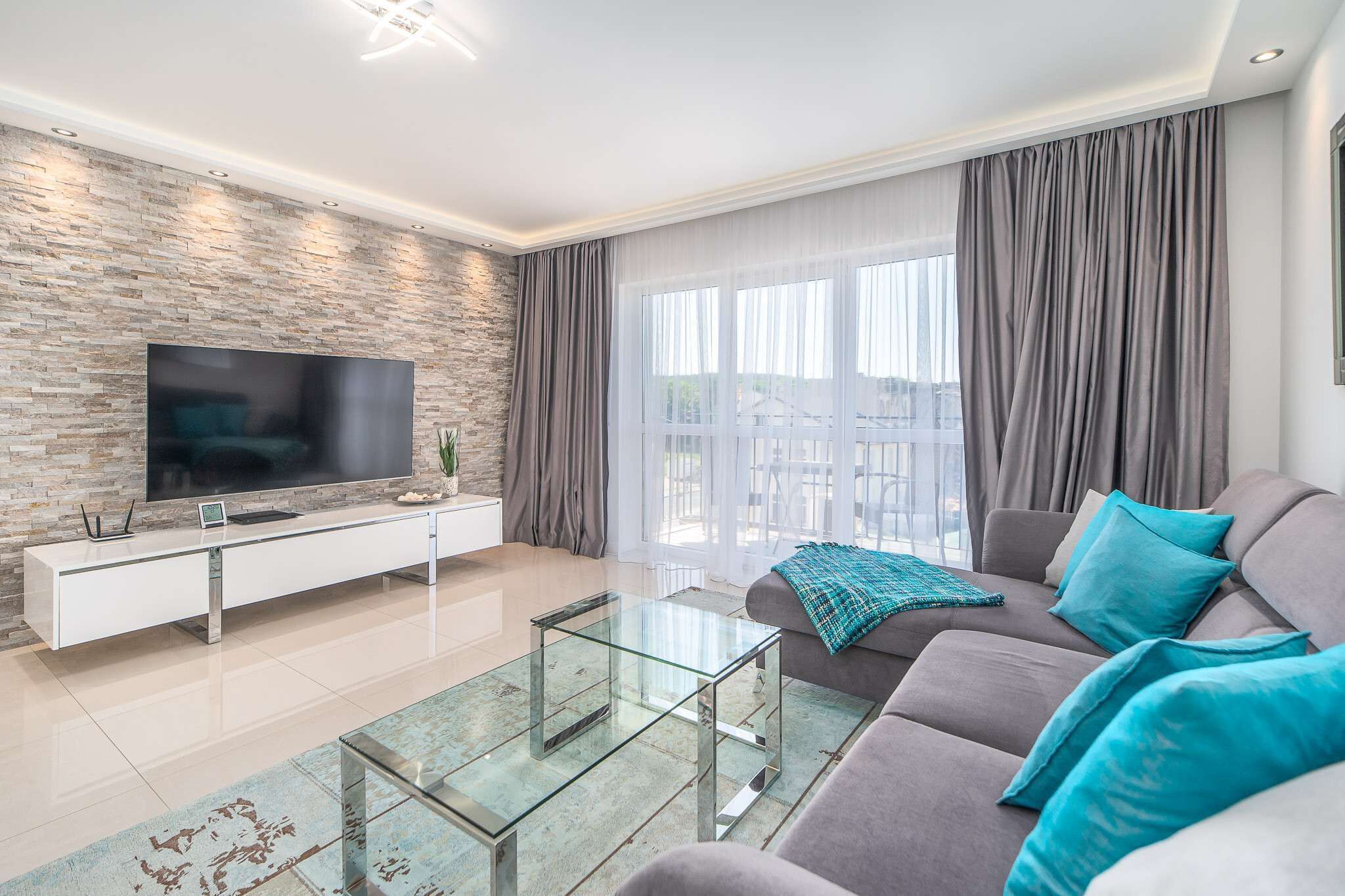 Un salon moderne avec un mur en pierre, un téléviseur à écran plat, une table basse en verre, un canapé gris et une grande fenêtre avec des rideaux.