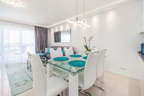 A modern living room with a glass dining table, white chairs, a sofa with blue cushions, a large mirror, and a potted plant.
