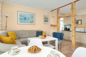 Un espacio de estar con un sofá, una mesa de comedor con un croissant y tazas de café, una silla azul y una cocina al fondo.