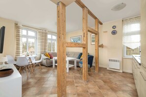 Un salon moderne avec un coin repas, des poutres en bois, un canapé blanc, une table basse et une cuisine dotée d’un radiateur blanc.